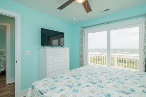 A bedroom with a bed, a TV on a dresser, and a view of the ocean through a sliding glass door.