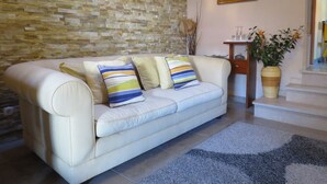 A living room with a beige sofa, striped pillows, a stone wall, a framed painting, a small table with a vase, and a staircase. - Azenhas do Mar Village - Holiday Rentals (Colares)