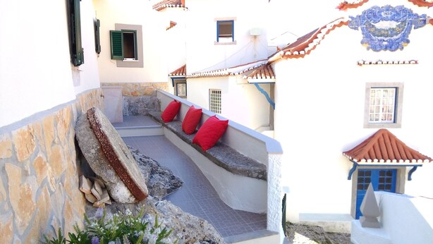 A narrow courtyard with stone steps leading to a seating area with red cushions, surrounded by white buildings with tiled roofs. - Azenhas do Mar Village - Holiday Rentals (Colares)