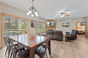 A spacious living room with a wooden dining table, comfortable seating, a fireplace, and large windows offering a view of the outdoors.