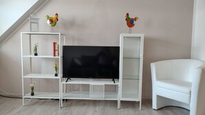 En stue med et fladskærms-tv på et hvidt stativ, to hvide reoler med pynteting og en hvid lænestol.
