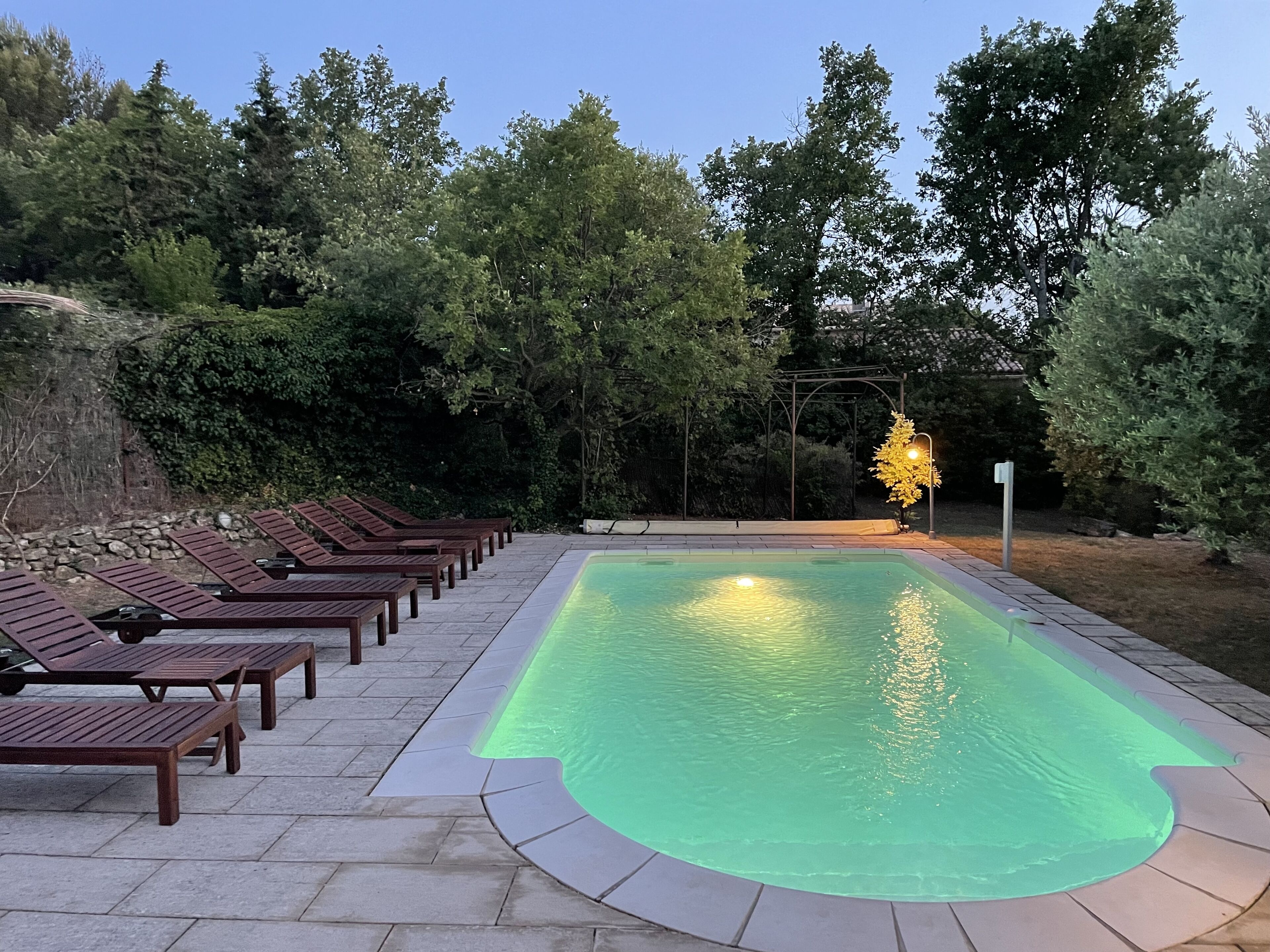 A swimming pool with a tiled deck and wooden lounge chairs, surrounded by trees and a stone wall.