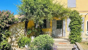Un camino de piedra que lleva a un edificio amarillo con puerta y ventanas, rodeado de diversas plantas y un muro de piedra.