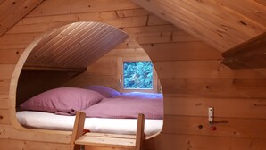 Ein Schlafzimmer im Loft-Stil mit holzverkleideter, schräger Decke, einem kleinen Fenster und einer Holzleiter, die zu einem Bett mit violetter Bettwäsche führt.