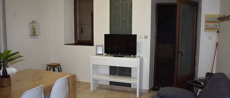 A living room with a TV on a white stand, a dining table with chairs, a gray sofa, and a sliding glass door.