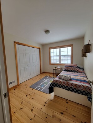 Ein Schlafzimmer mit Holzboden, einem Bett mit bunter Decke, einem Fenster und einer Tür.