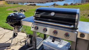A Weber grill with a digital thermometer, a side burner, and a built-in cooler, set up outdoors with a view of a lake and trees in the background. - Your Home Away From Home (Perth)