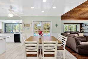 A modern kitchen with white cabinets, a wooden dining table, and a built-in oven. There is a large window with a view of the outdoors.