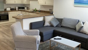 Un salon moderne avec un canapé gris, un fauteuil à carreaux et une petite table blanche. On aperçoit, au fond, une cuisine avec des meubles blancs et un four à micro-ondes.