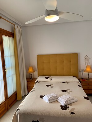 Un dormitorio con una cama, dos mesitas de noche con lámparas, un ventilador de techo, una ventana con cortinas y una puerta.
