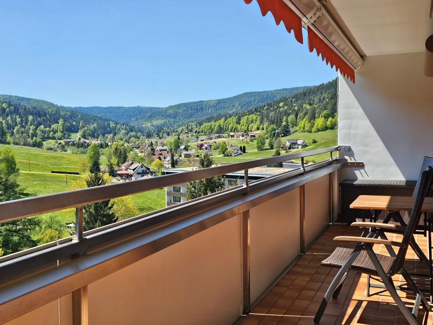 Ein Balkon mit Blick auf ein Dorf, ein Tisch mit Stühlen und ein rotes Sonnensegel.