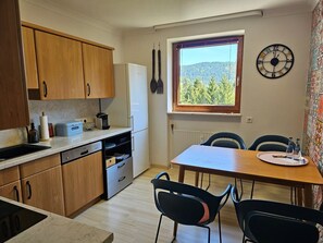Une cuisine de taille réduite, dotée de meubles en bois, d’un réfrigérateur blanc, d’une fenêtre donnant sur les arbres et d’un coin repas avec deux chaises et une table.