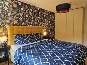 Une chambre à coucher avec un papier peint à motifs floraux, une tête de lit capitonnée, une couvre-lit à motifs, une lampe de chevet et une armoire.