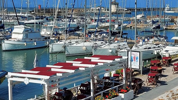 En lysthavn med mange både i havnen, et overdækket siddeområde med borde og stole, og en bygning i baggrunden.
