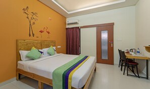 Une chambre d’hôtel avec un lit en bois, un bureau avec une chaise, un appareil de climatisation, une porte avec un panneau de verre et un mur orné d’une fresque représentant des oiseaux et un flamant rose.
