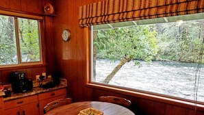 Ein Holzhäuschen-Interieur mit einem runden Esstisch, Stühlen, einer Kaffeemaschine und Blick auf einen Fluss durch große Fenster.