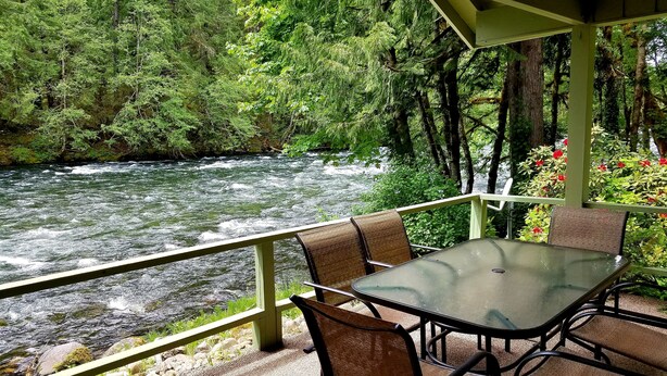 Ein überdachter Außenbereich mit einem Glastisch und Stühlen, mit Blick auf einen Fluss und einen dichten Wald.
