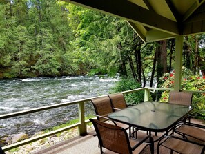 Un espace extérieur abrité, doté d’une table en verre et de chaises, offrant une vue sur une rivière sinueuse et une forêt dense.