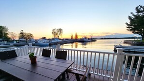 A wooden deck with a table and chairs, overlooking a marina with boats and a sunset.