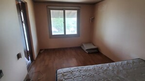 Une petite pièce vide, avec une fenêtre, un matelas et un plancher en bois.