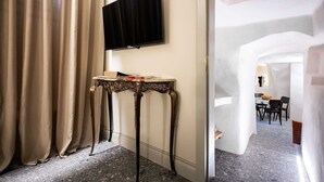 Una camera d'albergo con televisore a muro, un tavolino con un libro e vista su un'area ristoro attraverso un arco.