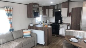 A compact kitchenette with a microwave, stove, and sink. There is a sofa with a striped pillow and a dining table with chairs. - Golden Palm Resort (Skegness)