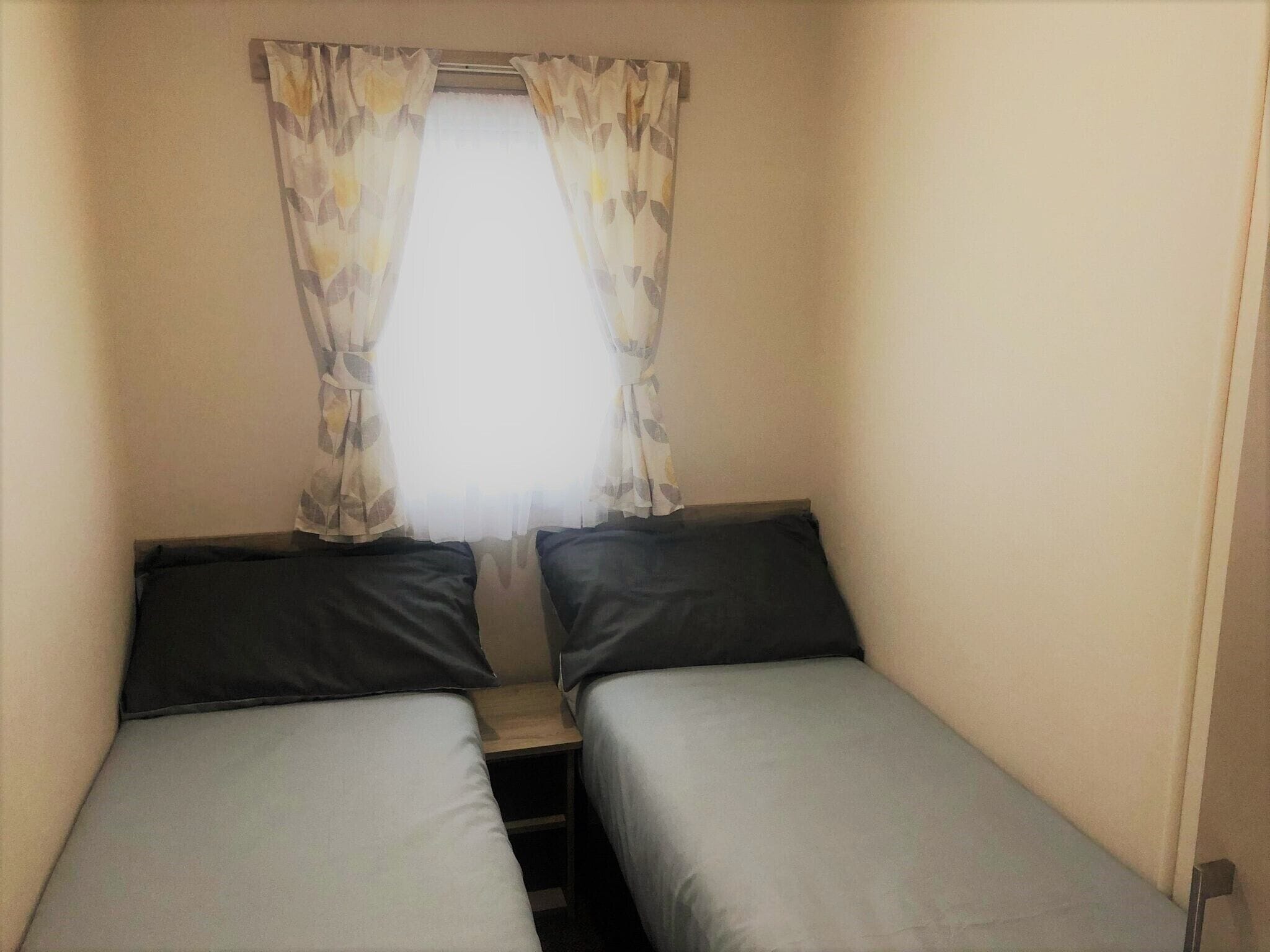 A small room with two single beds, a window with curtains, and a wooden nightstand.