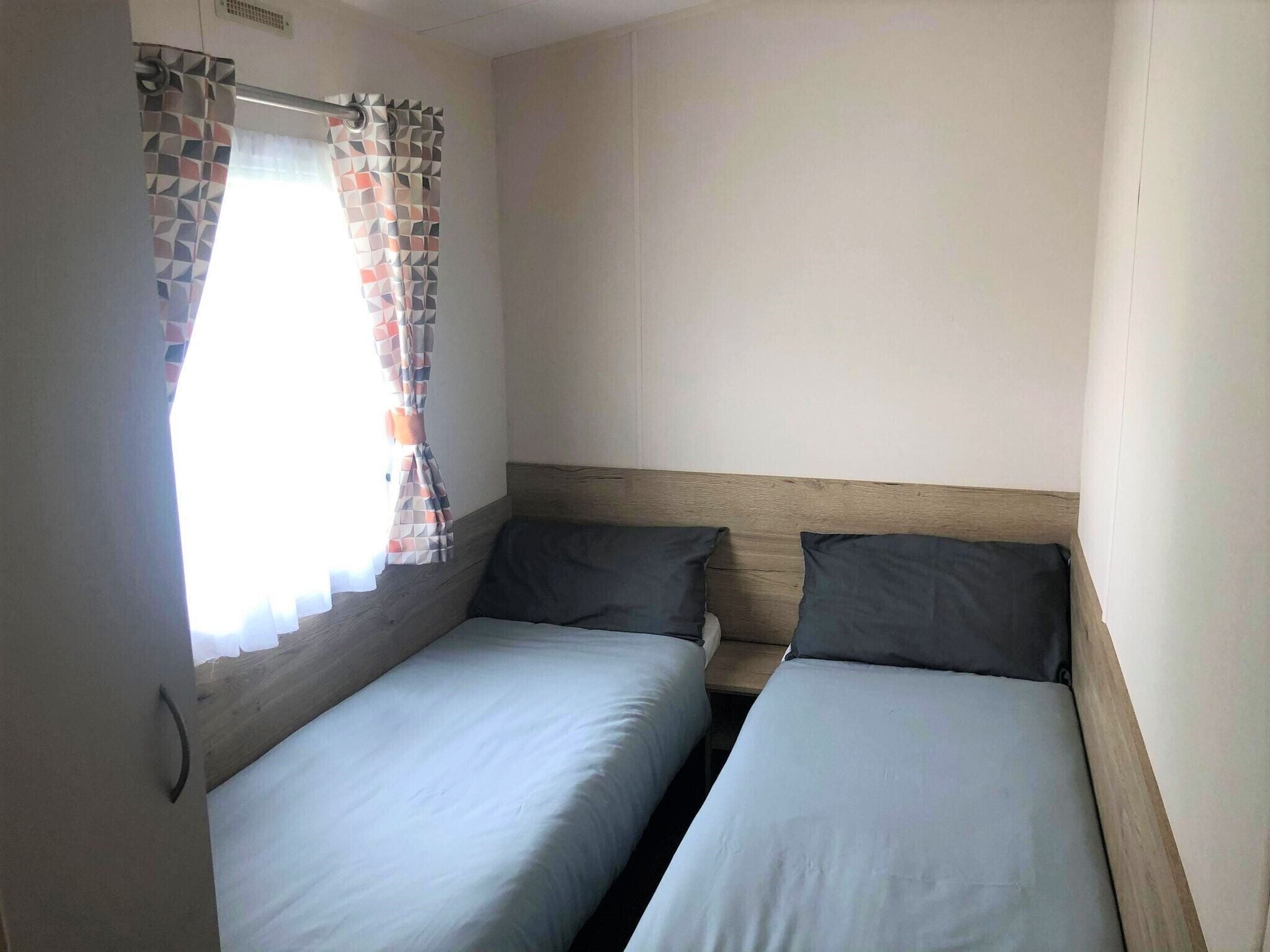 A small, single-room accommodation with two single beds, a window with a curtain, and a plain wall.