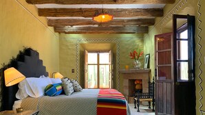 A bedroom with a large bed, a nightstand, a lamp, a fireplace, and a door leading to another room. - Meson Hidalgo (San Miguel de Allende)