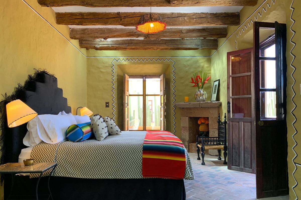 A bedroom with a large bed, a nightstand, a lamp, a fireplace, and a door leading to another room.