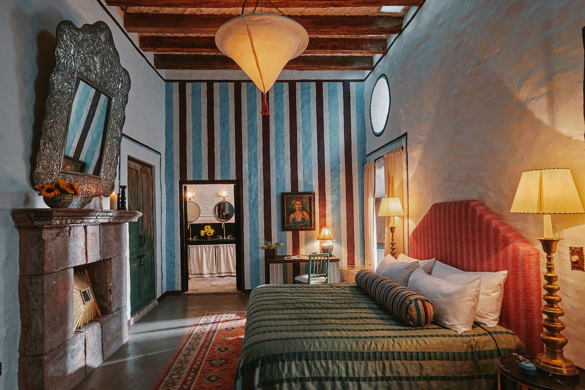 A room with a bed, a mirror, a fireplace, a desk, and a lamp.