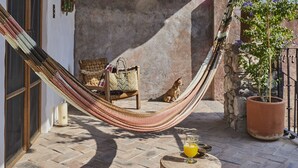 A hammock, wooden chairs, a wooden stool, and a potted plant in a courtyard with a stone wall and a hanging lantern. - Meson Hidalgo (San Miguel de Allende)