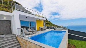 Un área de piscina moderna con vistas despejadas al océano, un muro de piedra y una escalera que conduce a un edificio con grandes ventanales.