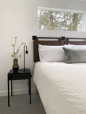 A bedroom with a bed, a nightstand, a vase with flowers, and a window with a view of trees.