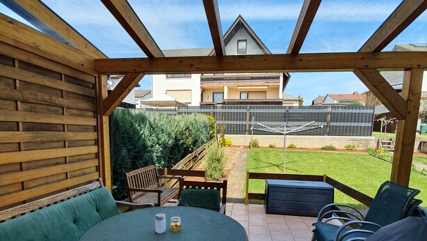 A covered patio with a wooden table and chairs, a green cushioned bench, and a view of a residential area. - Abgeschlossene Ferienwohnung (Altendiez)