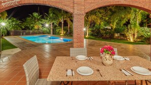 An outdoor dining area with a pool, brick arches, and tropical plants.