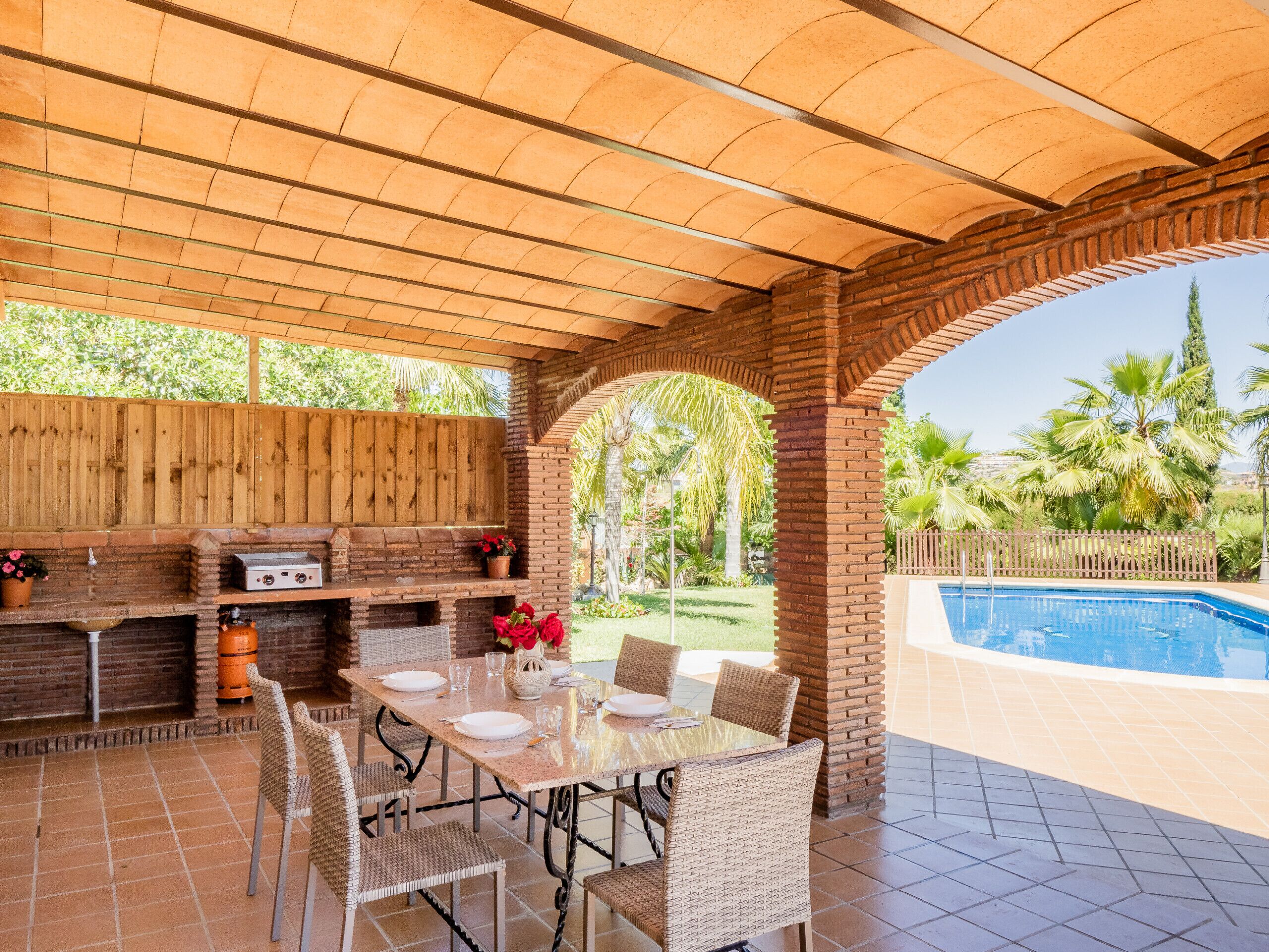 An outdoor patio with a brick barbecue, a pool, and a dining area with wicker chairs and a glass table.