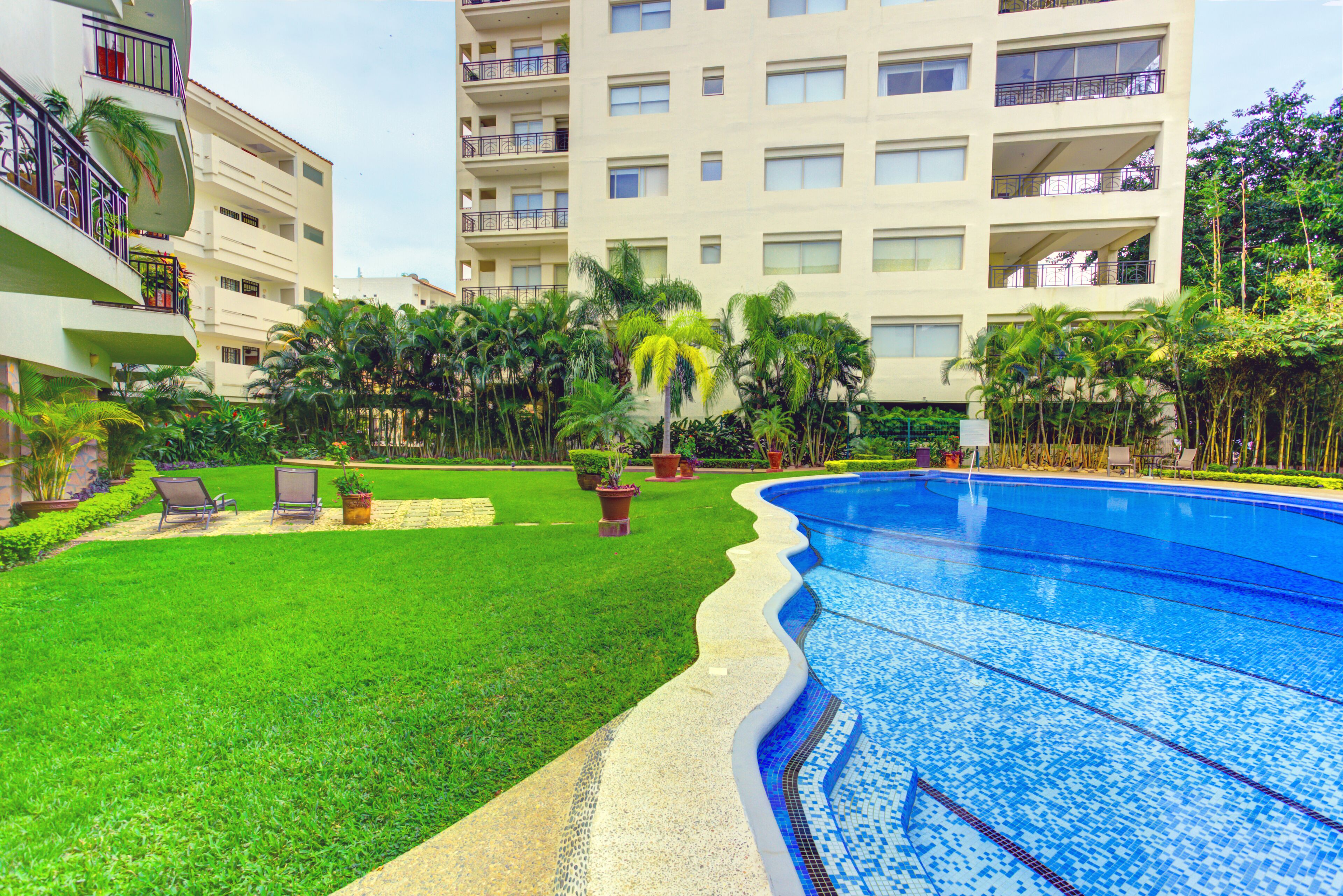 Una piscina rodeada de un césped cuidado, con un edificio de varios pisos al fondo.