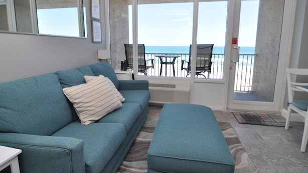 Una habitación con un sofá color turquesa, un reposapiés a juego, una mesita y una silla. Hay una puerta de cristal corredera que da a un balcón con vistas a la playa.