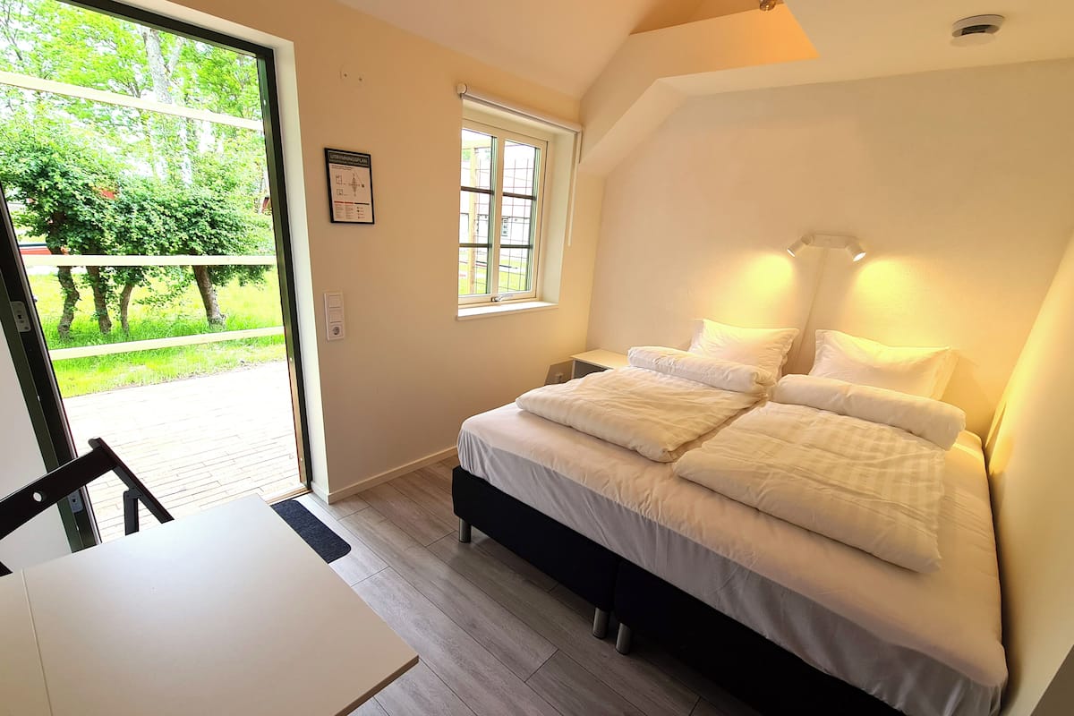 A bedroom with a bed, a window, a table, and a view of greenery outside.