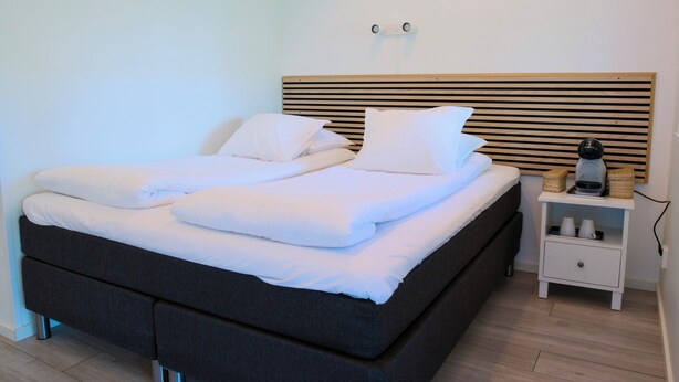 A neatly made bed with white linens and pillows, a wooden headboard, a bedside table with a coffee maker, and a drawer. - GEG Hotel (Landskrona)