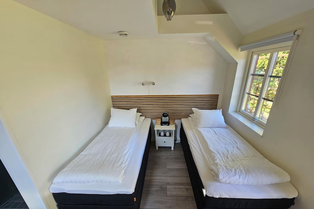 A small, clean room with two single beds, a bedside table with a lamp, and a window with a view of trees.