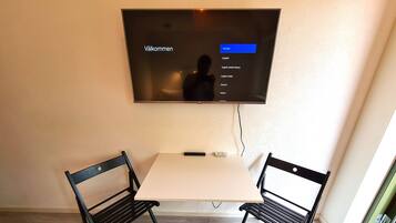 Ein Zimmer mit an der Wand befestigtem Fernseher, der ein Menü zur Sprachauswahl anzeigt. Es gibt einen kleinen Tisch mit zwei Stühlen, eine Fernbedienung auf dem Tisch und eine Tür mit grünem Rahmen rechts.