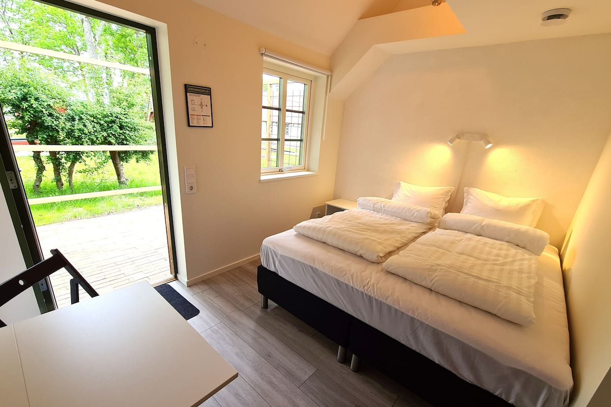 A bedroom with a bed, a window, a table, and a view of greenery outside.