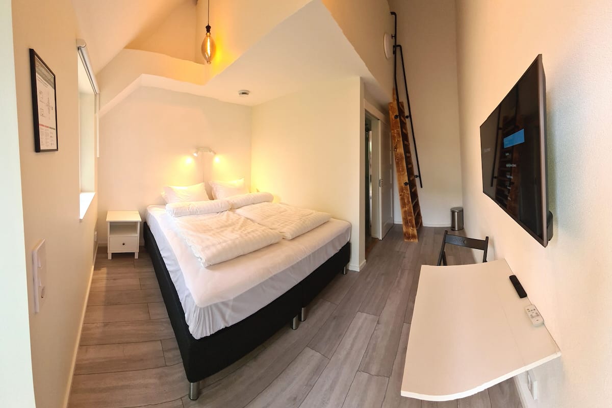 A bedroom with a bed, bedside tables, a desk, a chair, a TV, and a wooden floor.