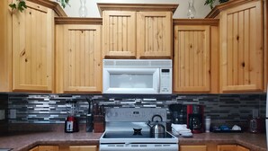 A kitchen with wooden cabinets, a white microwave, a stove, and a tiled backsplash. - I Deal Lake Powell Home 3BR, Jacuzzi, BBQ & Firepit (Page)