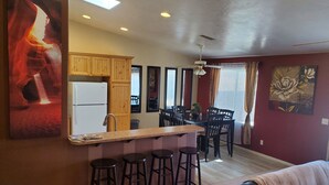 A modern kitchen with wooden cabinets, a white refrigerator, and a bar area with stools. There is a dining table with chairs in the background. - I Deal Lake Powell Home 3BR, Jacuzzi, BBQ & Firepit (Page)