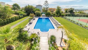 Et rektangulært swimmingpool med tilhørende træterrasse, tennisbane og varieret beplantning.