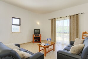 A living room with a sofa, a coffee table, a television on a stand, and a window with curtains.