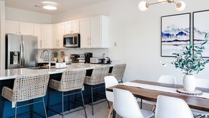 A modern kitchen with white cabinets, stainless steel appliances, and a dining area with wicker chairs and a wooden table. - ⭐️ Spacious Mountaintop Oasis at Camelback | 2,500 SF w/ Game Room, Deck & A/C (Tannersville)